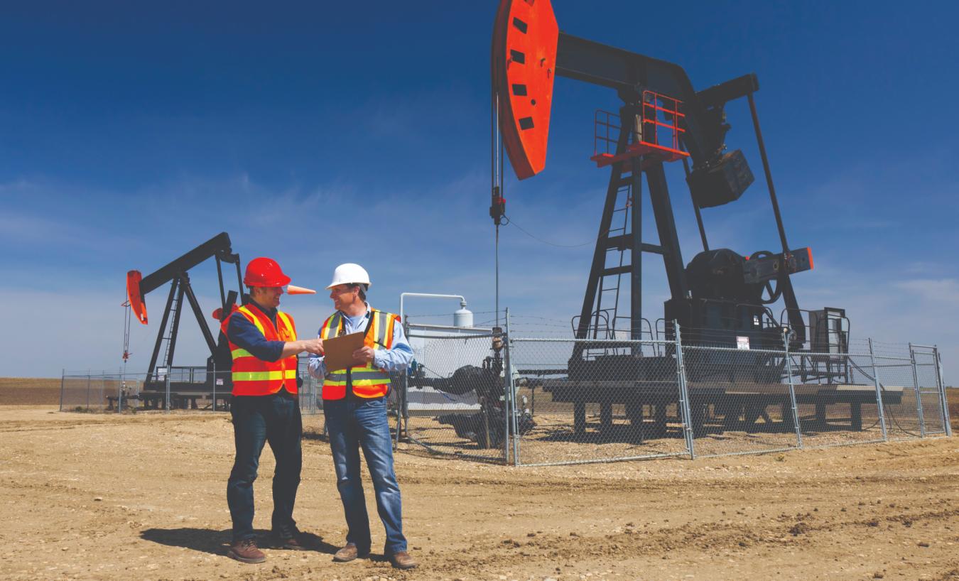 Two workers in hard hats and hi-vis vests reviewing a clipboard beside pump jacks at an oil field