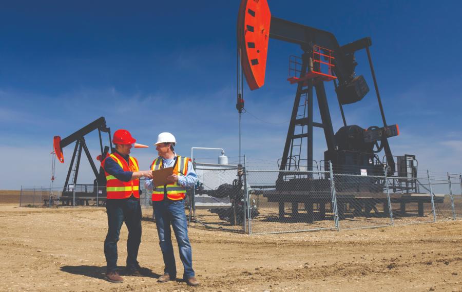 Two workers in hard hats and hi-vis vests reviewing a clipboard beside pump jacks at an oil field