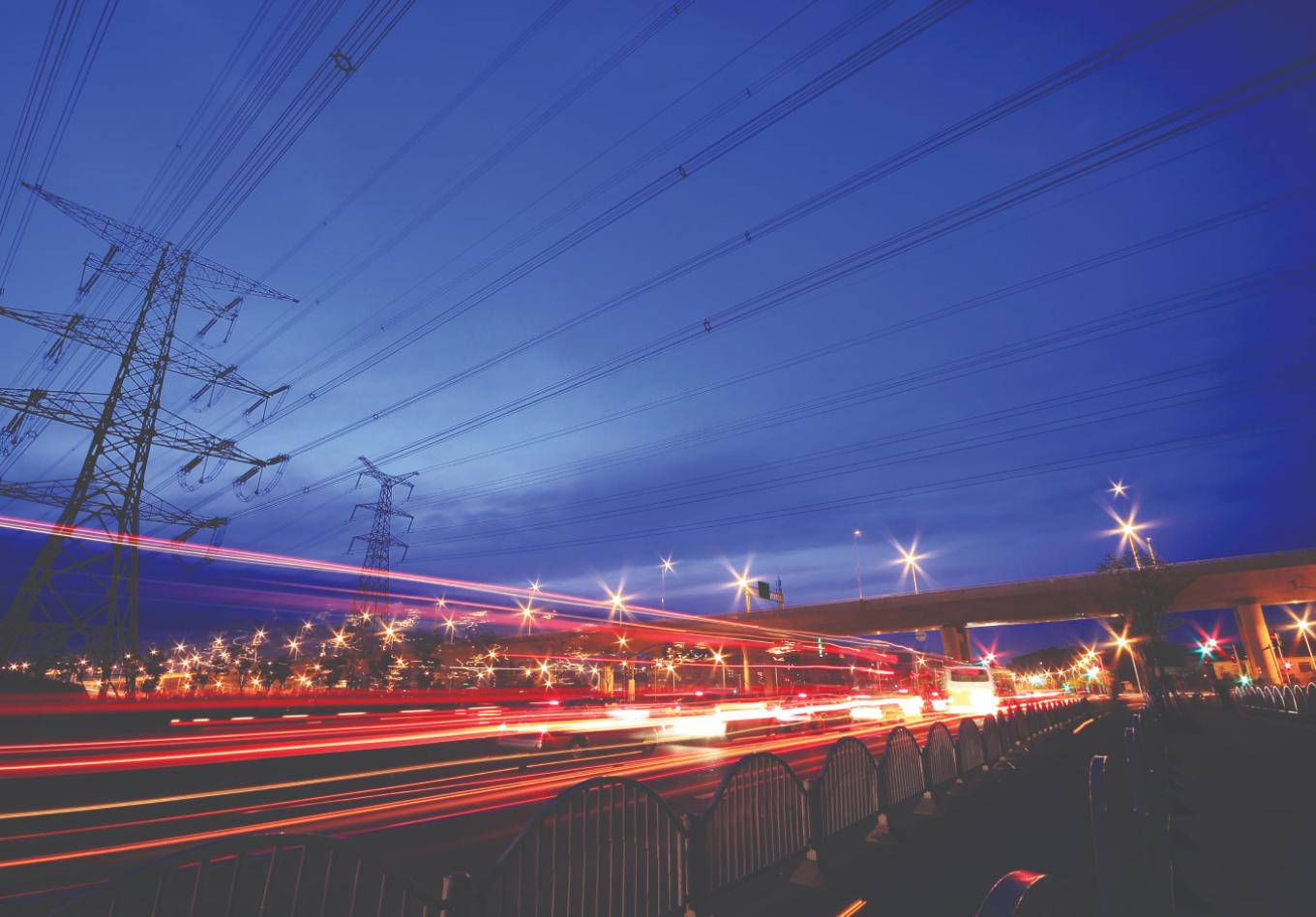 Electricity transmission pylons and power lines above a busy road at dusk, with light trails from passing traffic